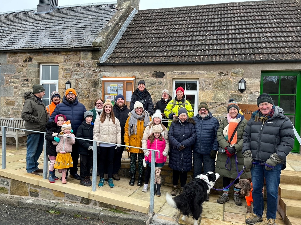 Group of adults, children and two dogs in outdoor gear outside community centre